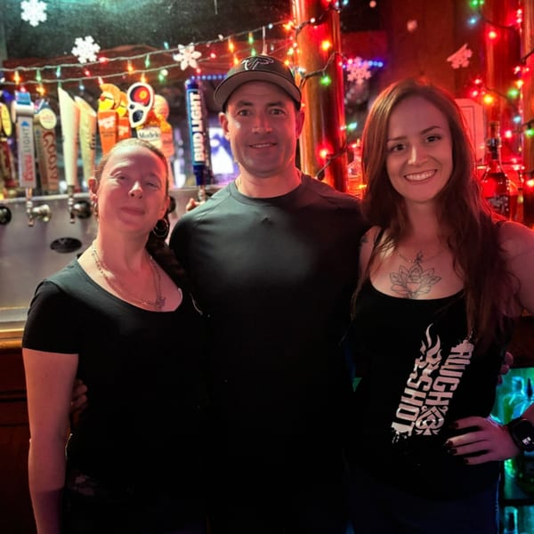Three smiling people, two women and one man, posing in front of a bar decorated with festive lights.