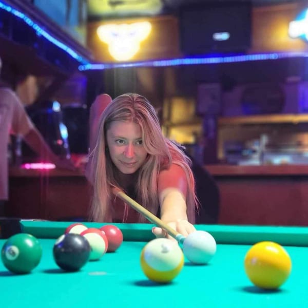 A woman with long blonde hair aims a pool cue at colorful billiard balls on a green table.