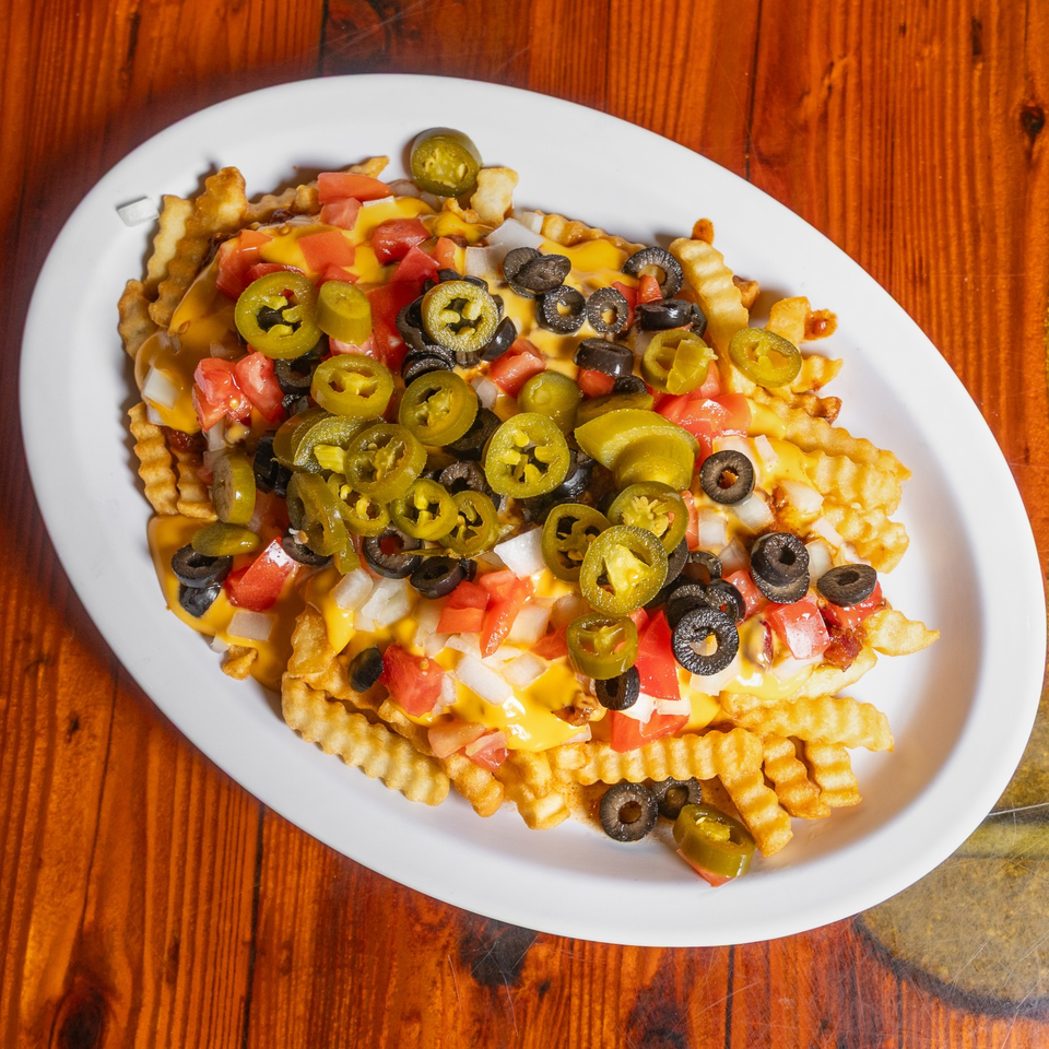 Loaded plate of crinkle-cut fries topped with melted cheese, jalapeños, black olives, tomatoes, and onions on a wooden table.