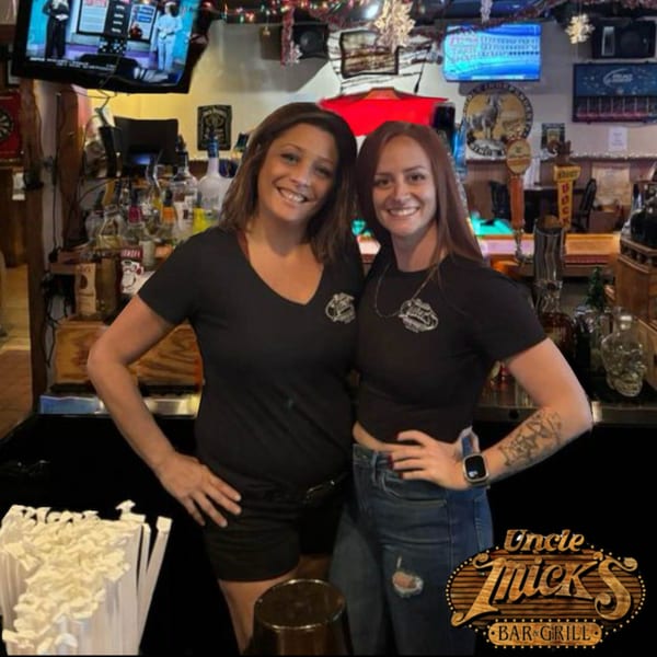 Two smiling women in black Uncle Mick's Bar & Grill shirts stand behind a bar.
