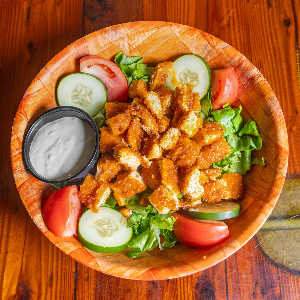 Crispy sauced chicken pieces served over lettuce with sliced cucumbers, tomatoes, and creamy dipping sauce on a wooden platter.