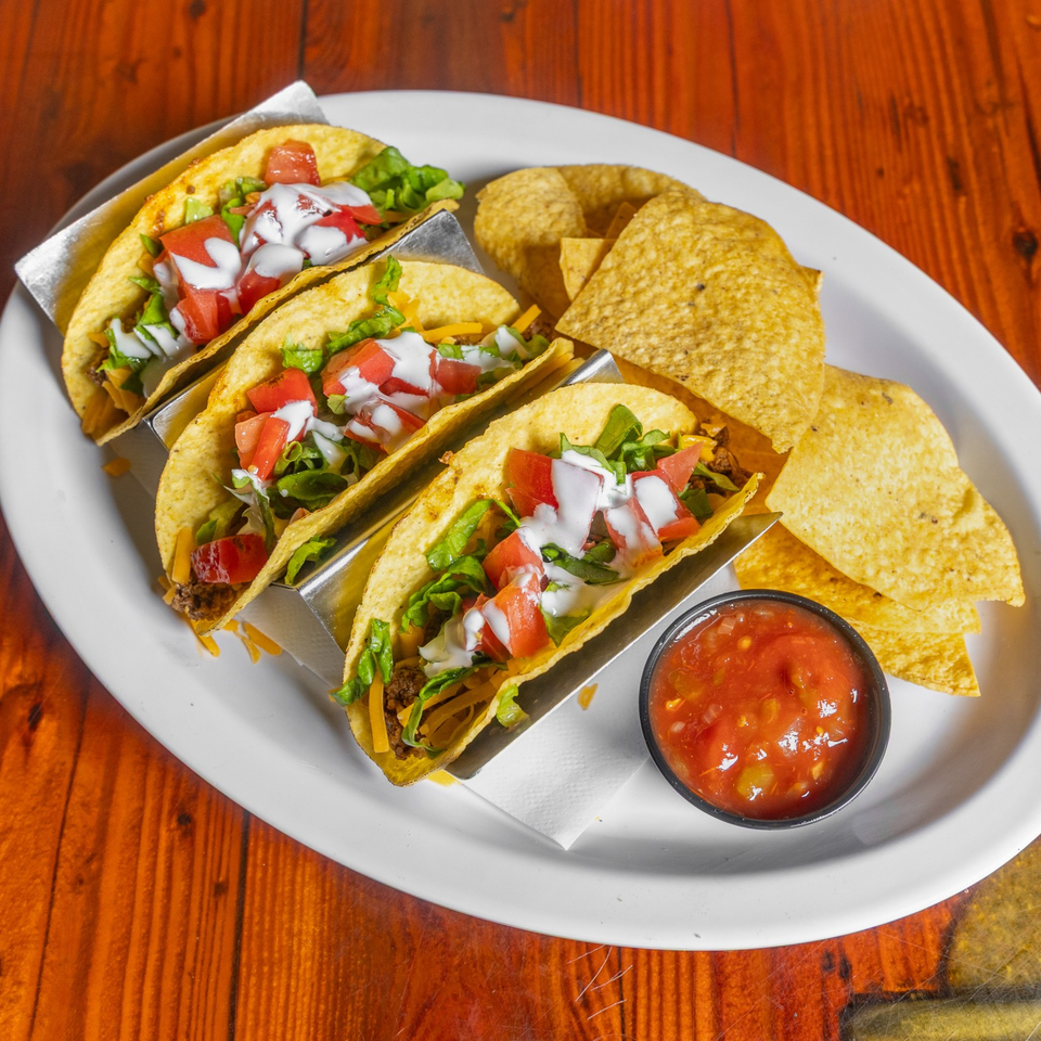 Three beef tacos topped with lettuce, tomatoes, cheese, and sour cream, served with tortilla chips and salsa on a plate.