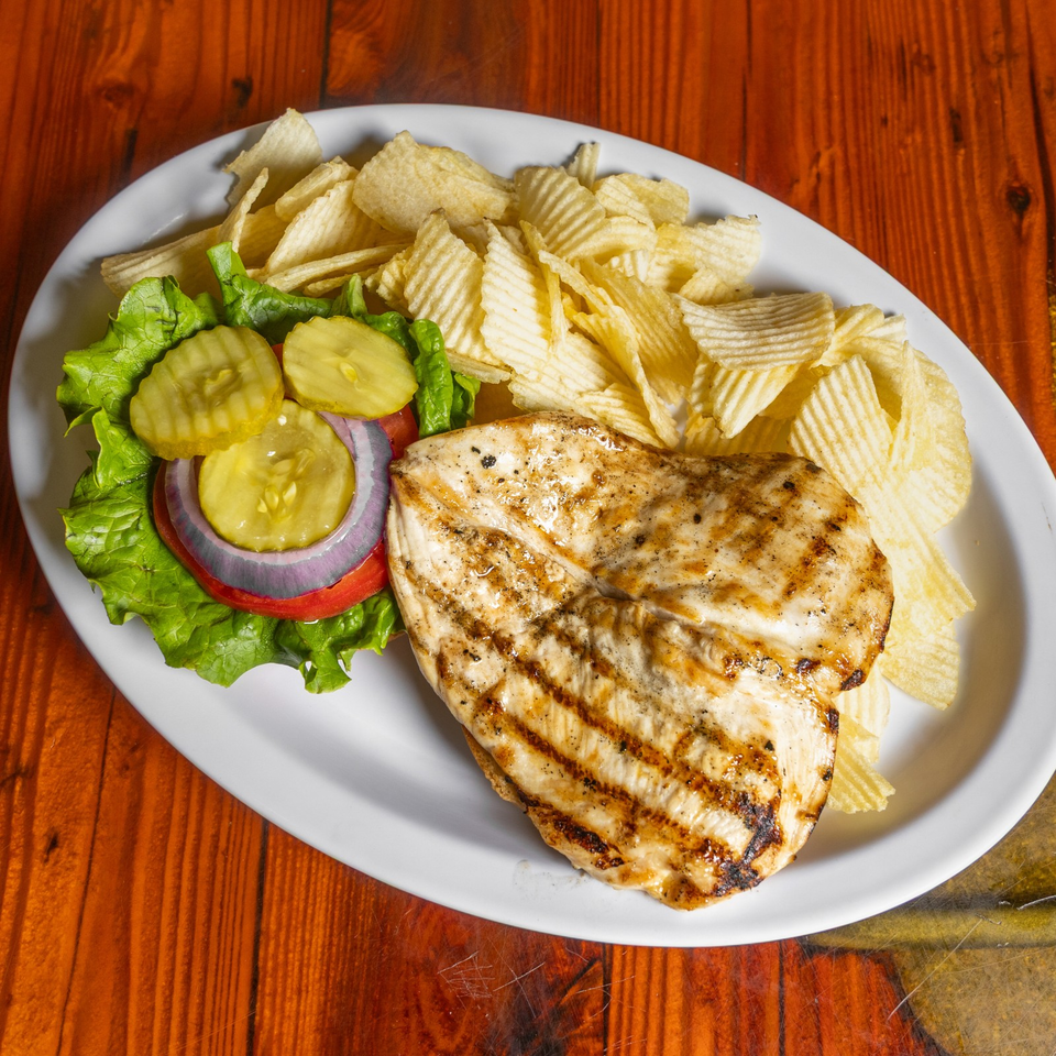 Grilled chicken sandwich with lettuce, tomato, onion, pickles, and ridged potato chips on a white plate.