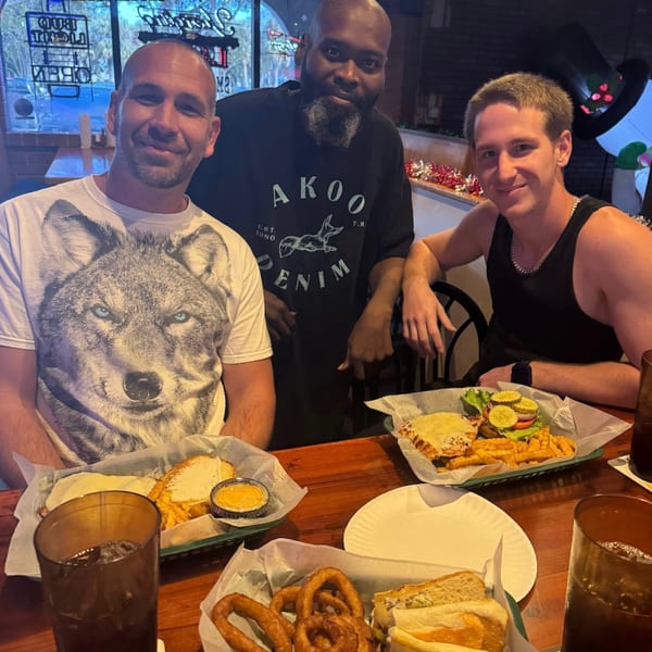 Three men smiling at the camera, seated at a table with burgers, fries, and onion rings.