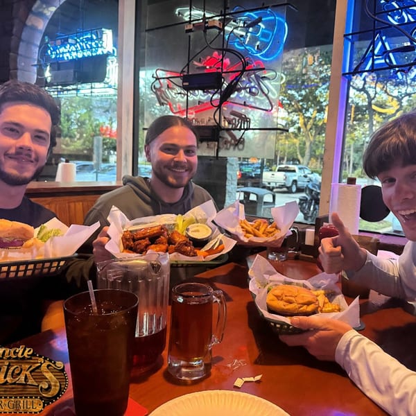 Three smiling men at Uncle Mick's Bar & Grill, enjoying wings, burgers, and fries.
