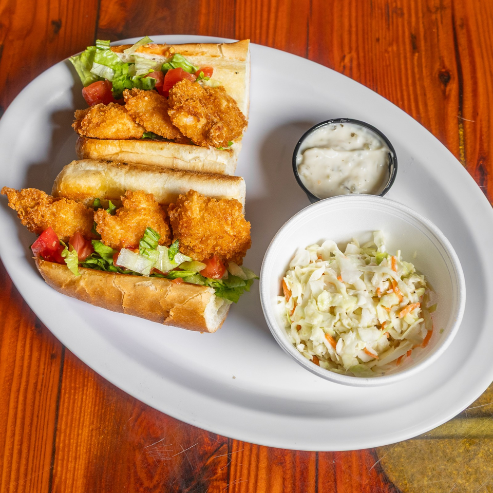 Two fried shrimp sandwiches with lettuce and tomatoes, served with coleslaw and creamy dipping sauce on a white plate.