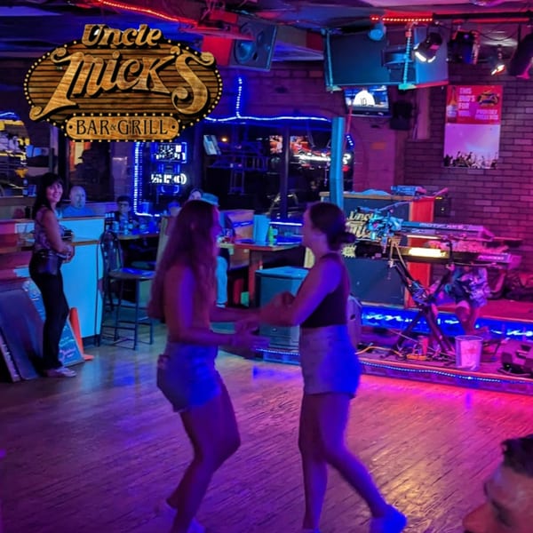 Two women dancing on a wooden floor at Uncle Mick's Bar & Grill, illuminated by blue and pink lights.